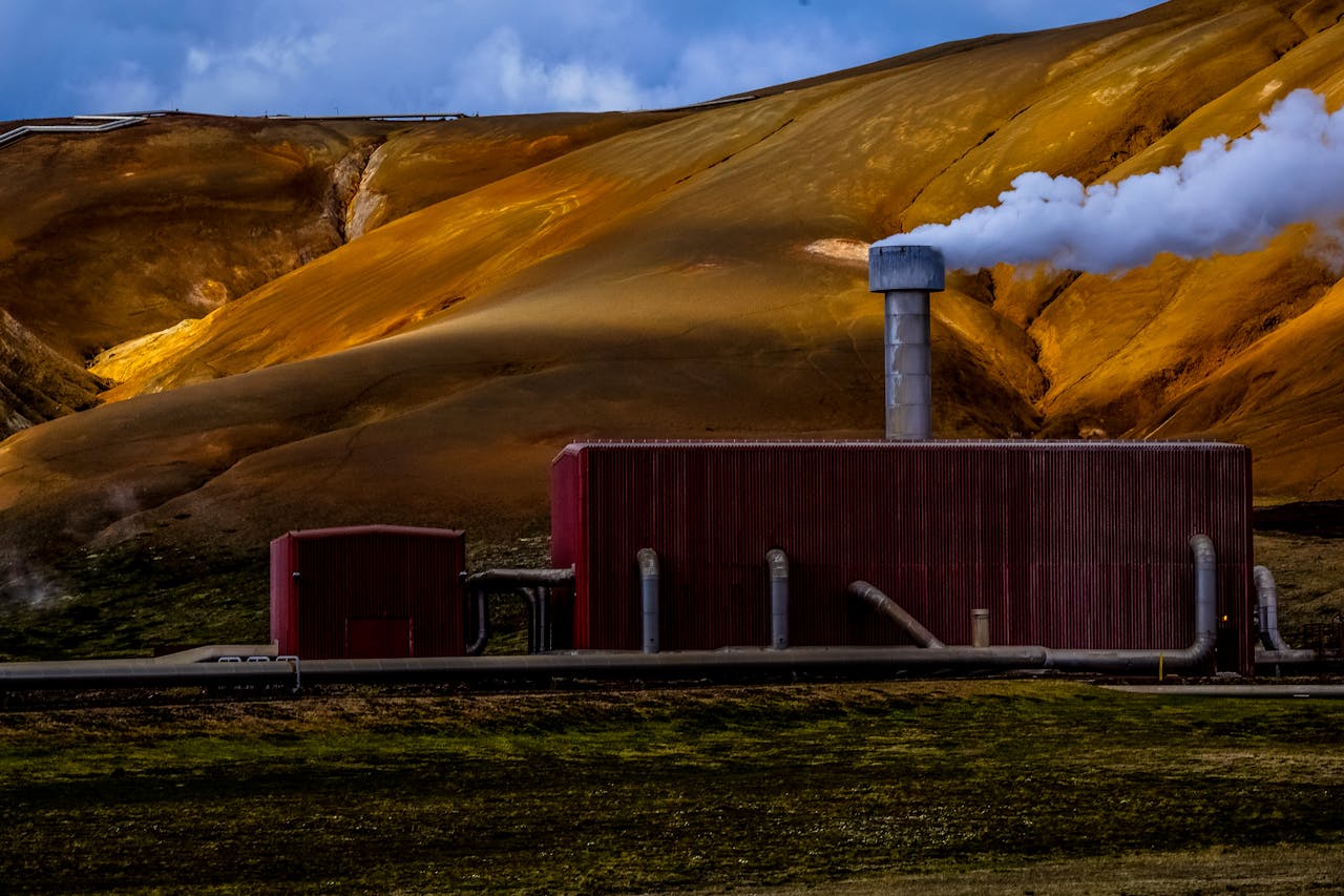 Red geothermal power plant emitting steam against a dramatic yellow-brown mountainous backdrop.